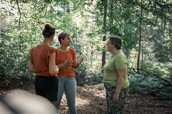 Drie vrouwen, waaronder Ellen Baas, in gesprek tijdens een wandeling in het bos, omringd door groen en zonlicht tussen de bomen.